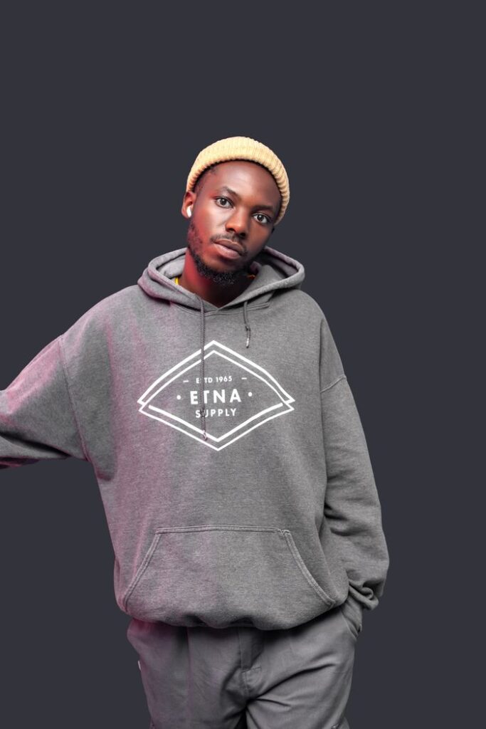 Stylish man wearing a grey hoodie and beanie, looking relaxed against a dark background.