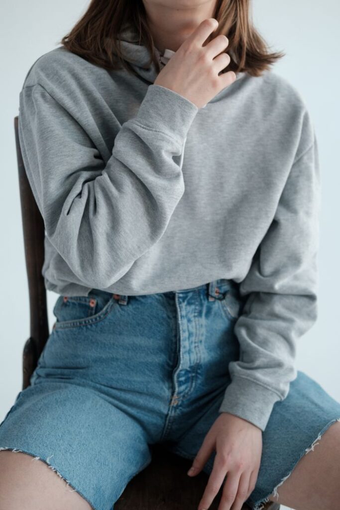 Anonymous slender female model wearing casual gray hoodie and denim shorts sitting on wooden chair on white background in studio
